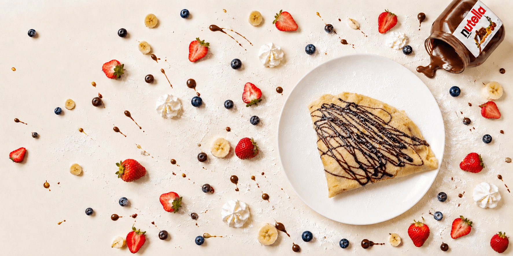 A fresh Nate's Crepes crepe on a plate, surrounded by strawberries, blueberries, banana slices, whipped cream, chocolate drizzle and a jar of Nutella