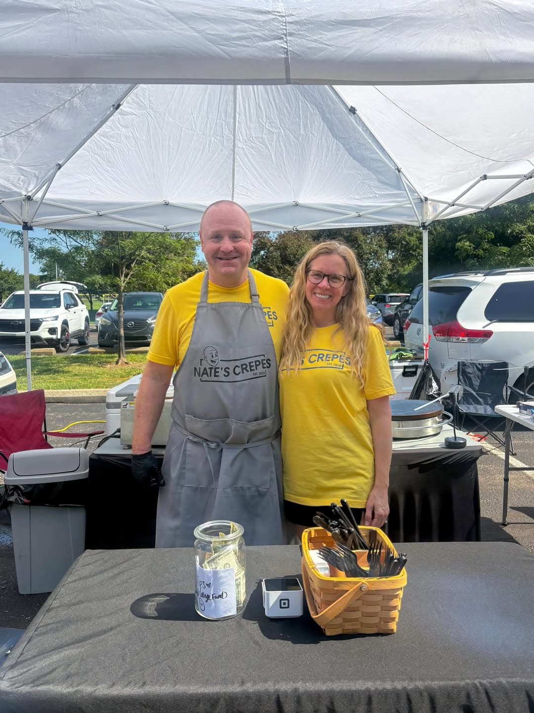 Nate and Carla standing together at their Nate's Crepes market stand, both wearing matching yellow Nate's Crepes t-shirts under their tent